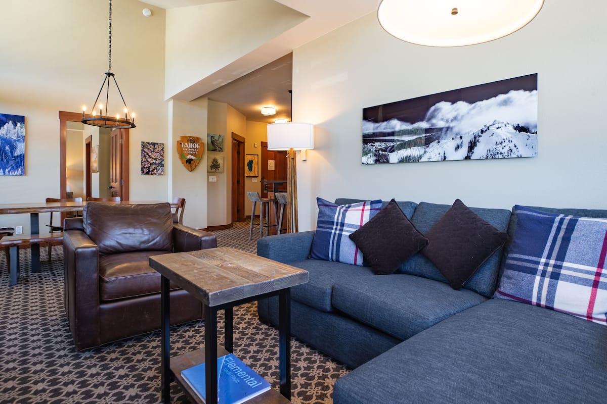 A modern living room with a leather chair, a sofa with plaid cushions, a wooden coffee table, and a large painting of a mountain landscape.