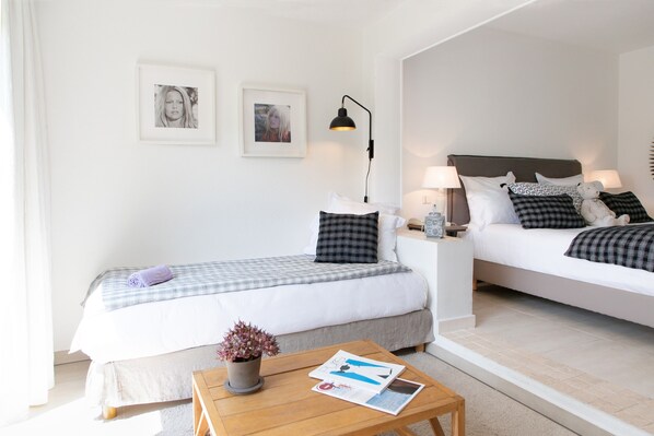 A hotel room with two beds, a bedside table with lamps, a small table with a plant and magazines, and framed pictures on the wall.