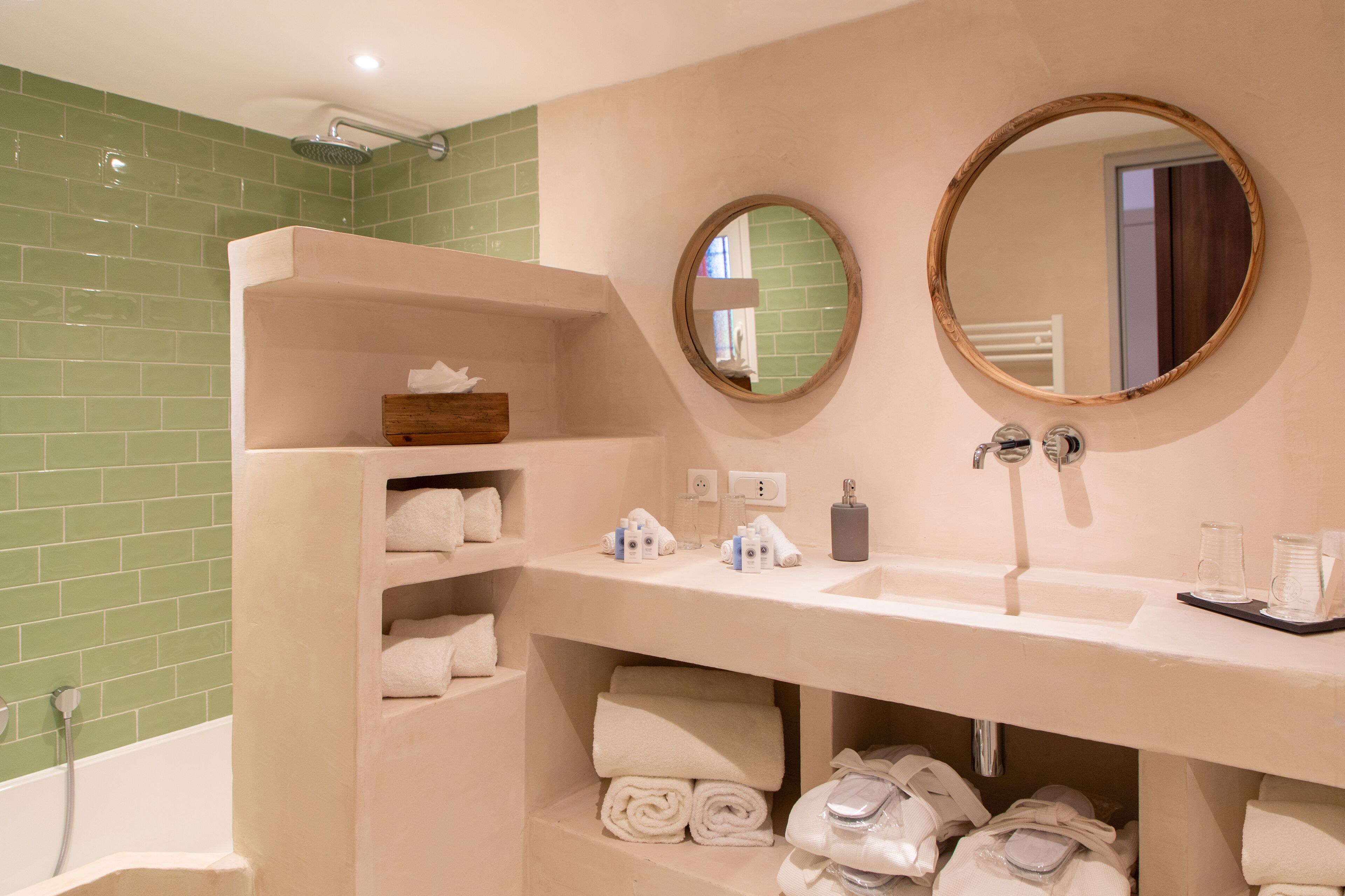 Une salle de bain avec un meuble-lavabo beige, deux miroirs ronds, une douche carrelée de carreaux verts et des serviettes soigneusement pliées.