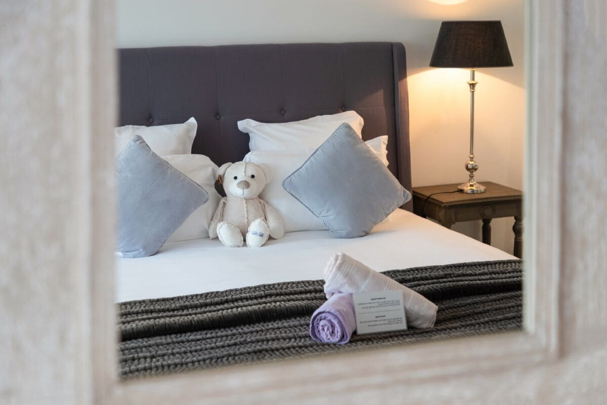 Una cama bien hecha con almohadas, un osito de peluche y una toalla enrollada sobre un tapete a rayas.