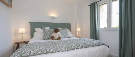A neatly made bed with a teddy bear, two bedside lamps, and a window with curtains.