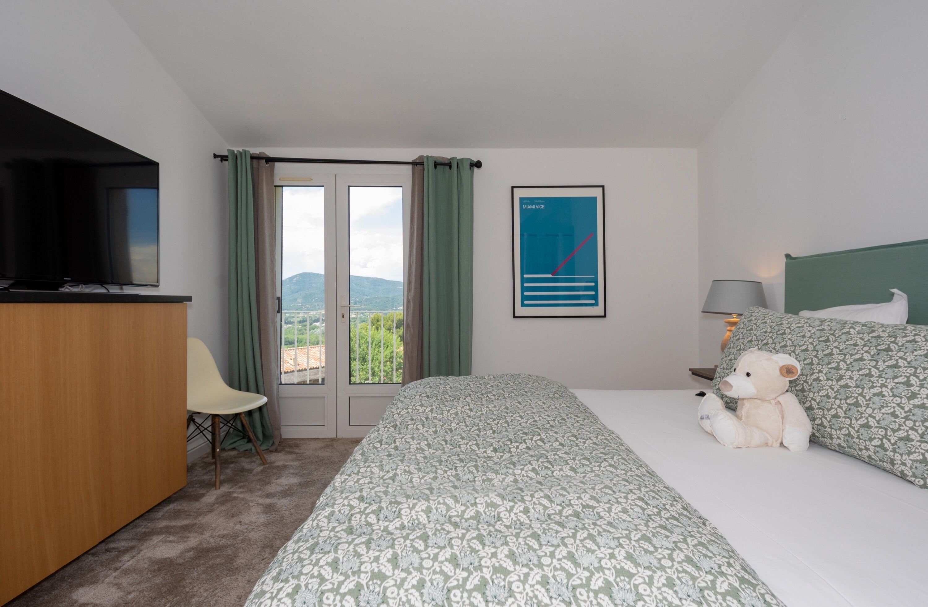 Een hotelkamer met een bed, een stoel, een televisie, een raam met gordijnen en een poster aan de muur.