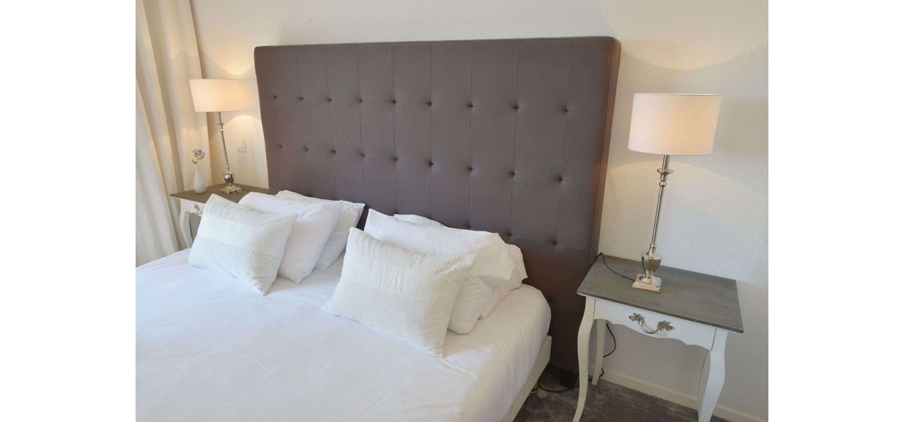 Een net opgemaakt bed met een gestuwd hoofdbord, twee nachtkastjes met lampen, en een effen muur.