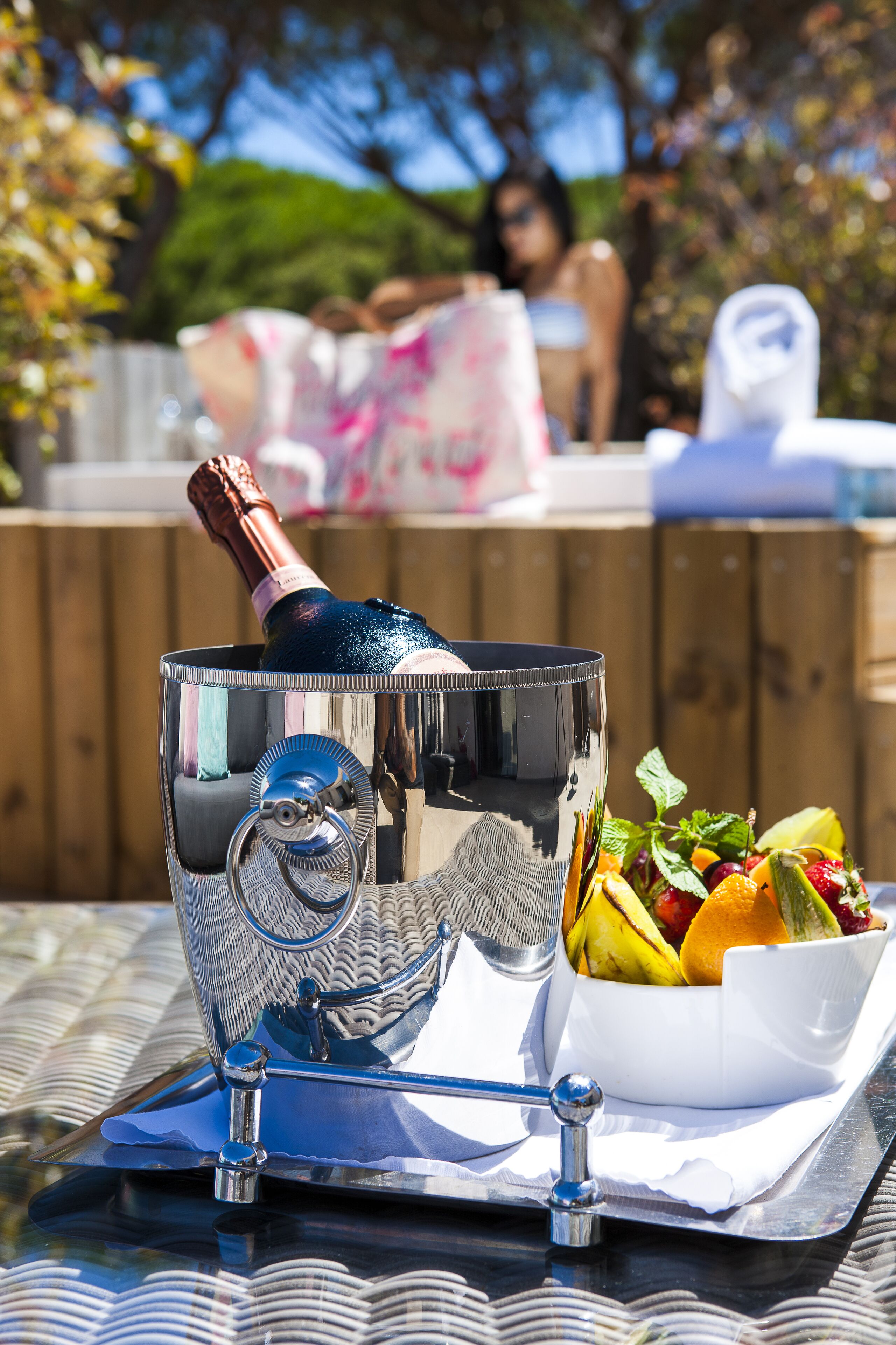 A metallic champagne bucket with a bottle, a bowl of fruit, and a napkin on a table outdoors.