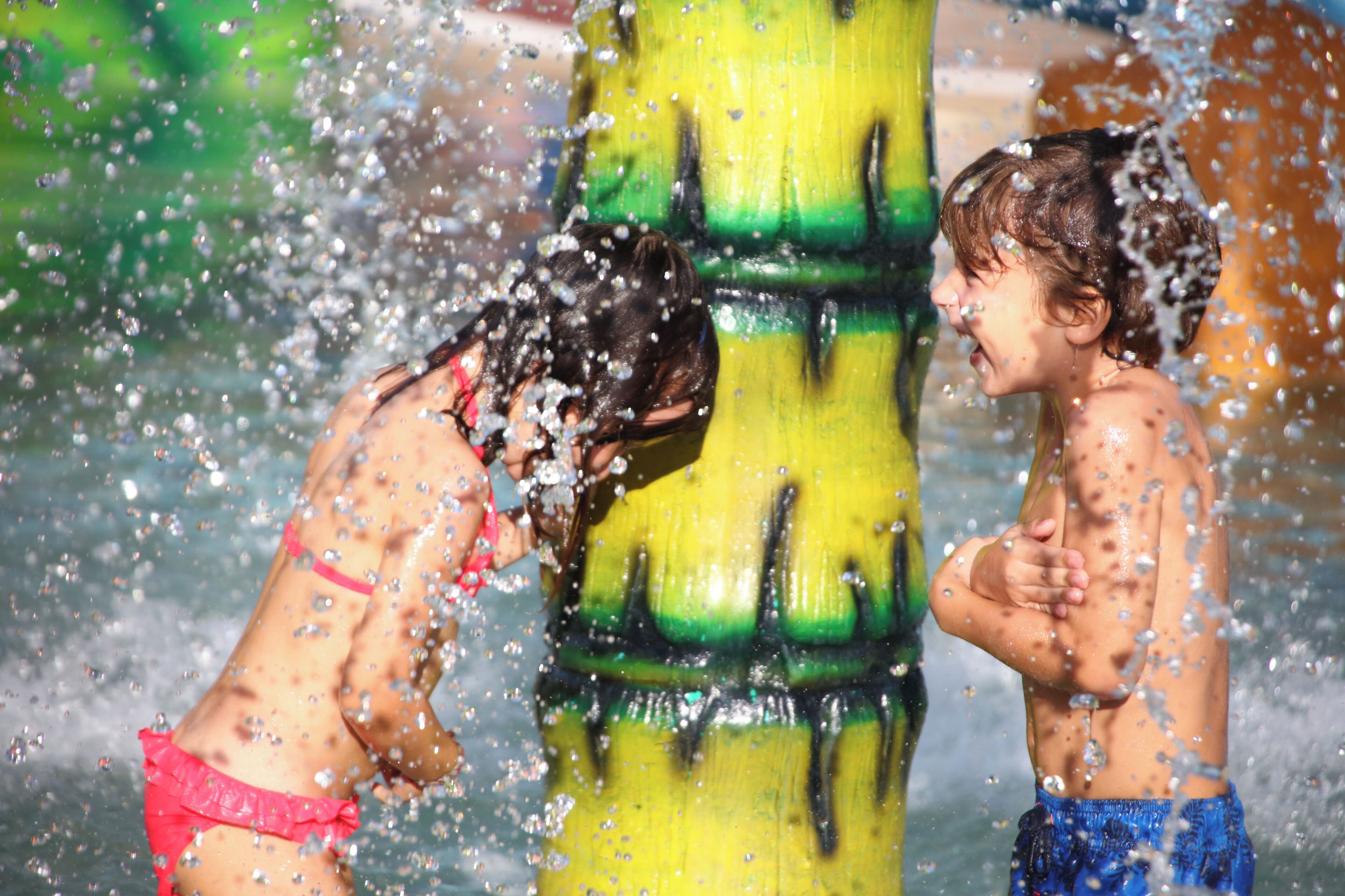 Two children playing in water, with a large yellow and green structure in the background.