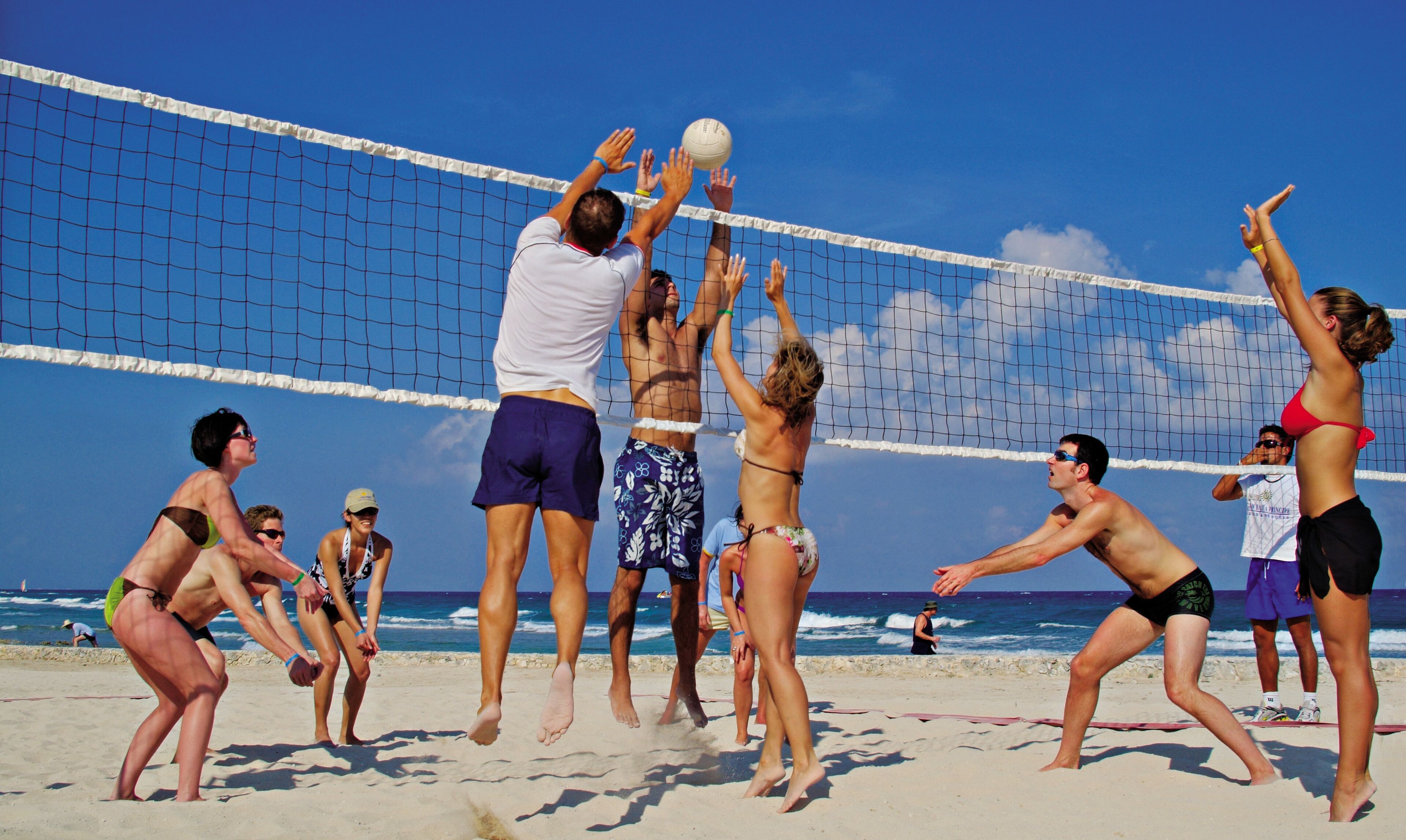 A beach volleyball game with players in swimwear, a clear sky, and the ocean in the background.