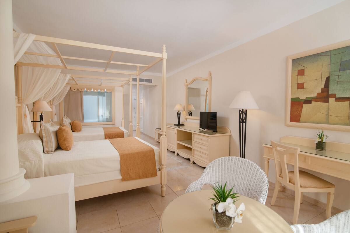 A hotel room with a four-poster bed, a desk with a computer, a chair, and a dining table with a plant.