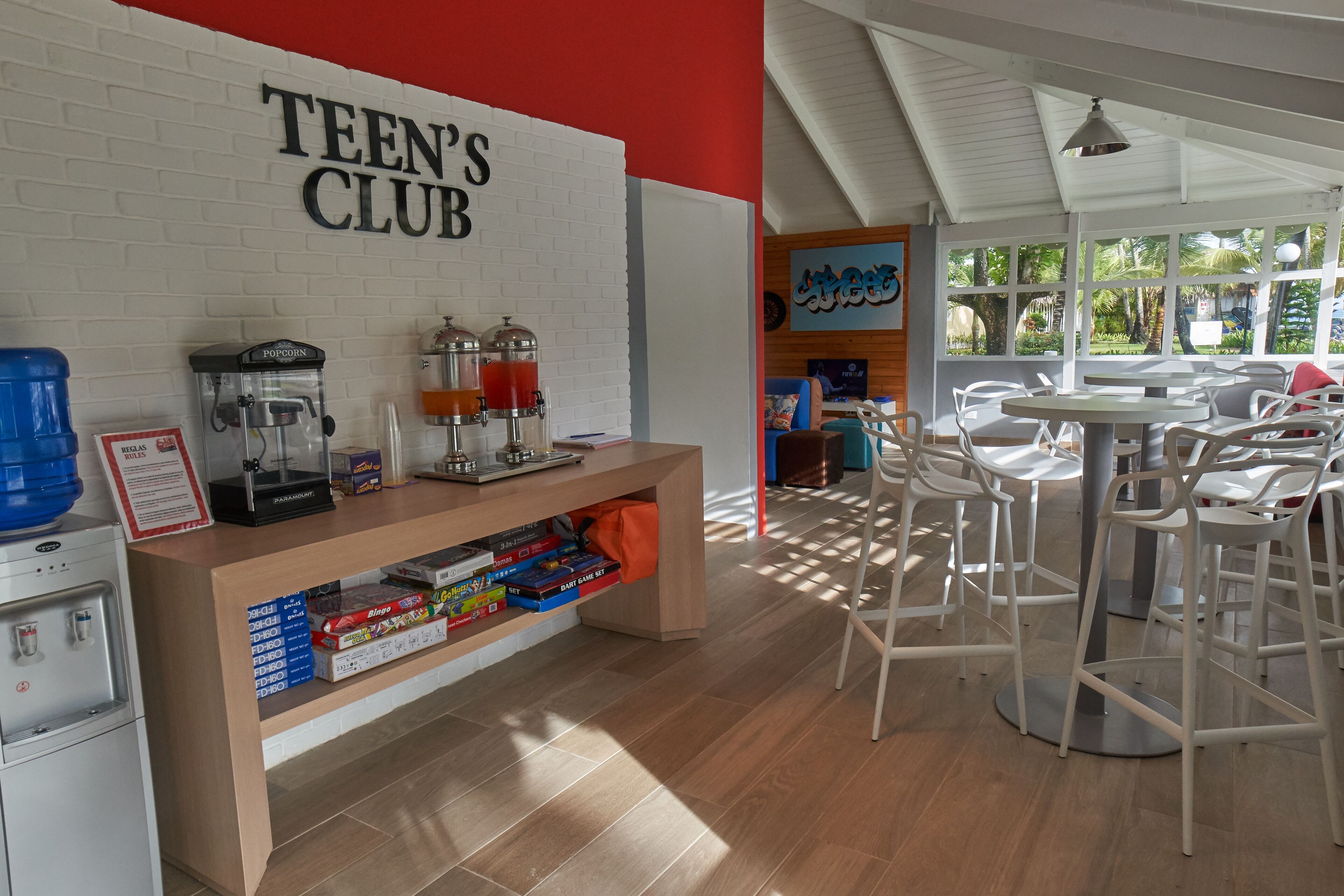 Jugendclub mit weißer Backsteinwand, einer Popcornmaschine, einem Getränkespender und einer Theke mit Gesellschaftsspielen. Weiße Stühle und Tische sind zum Sitzen bereitgestellt.