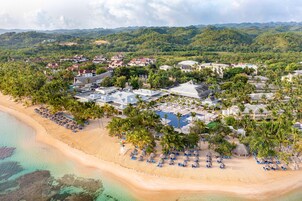 Vista aérea de un resort con propiedades frente a la playa, una piscina y palmeras.