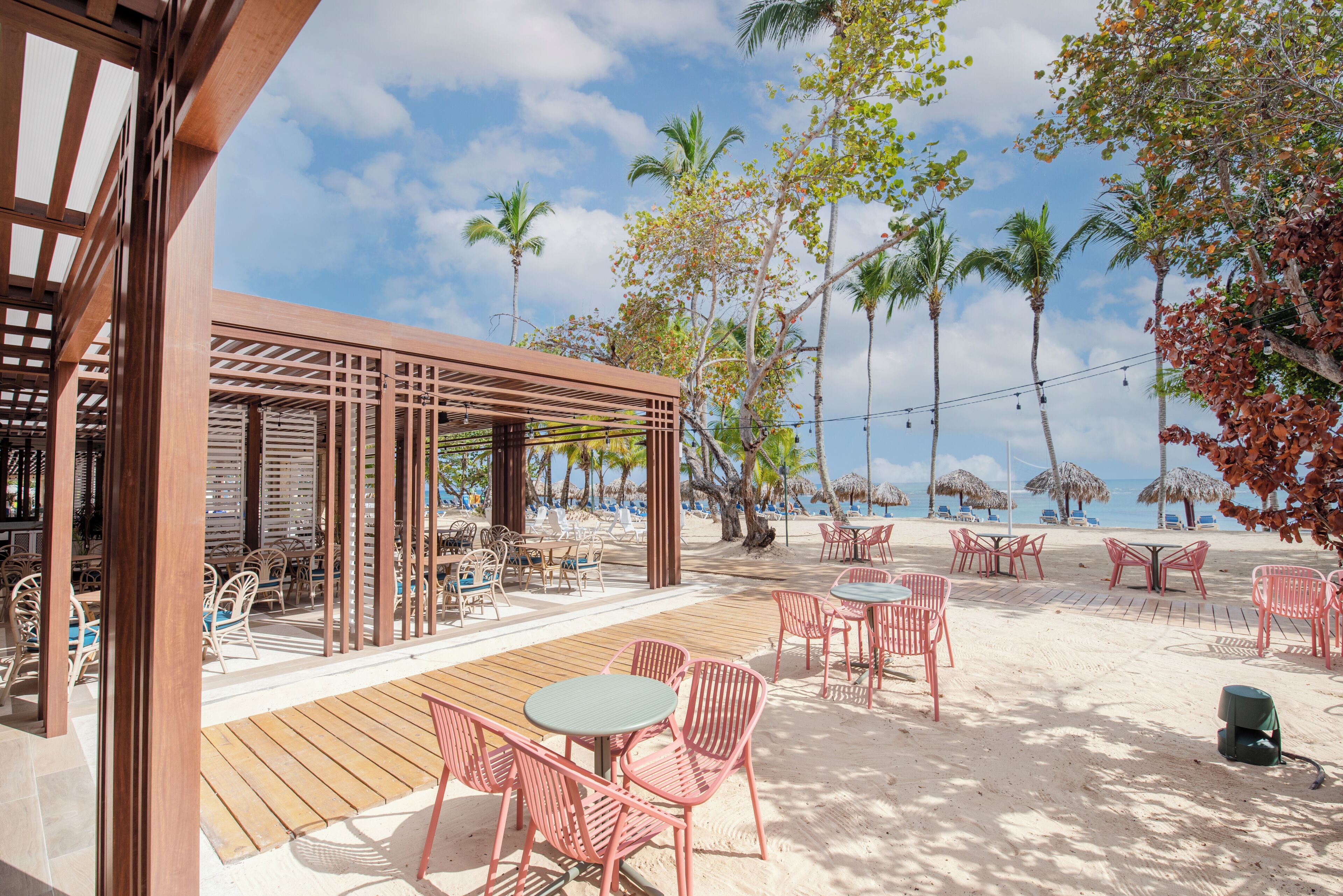 A beachside patio with wooden flooring, red metal chairs, and tables, surrounded by palm trees and beach umbrellas.