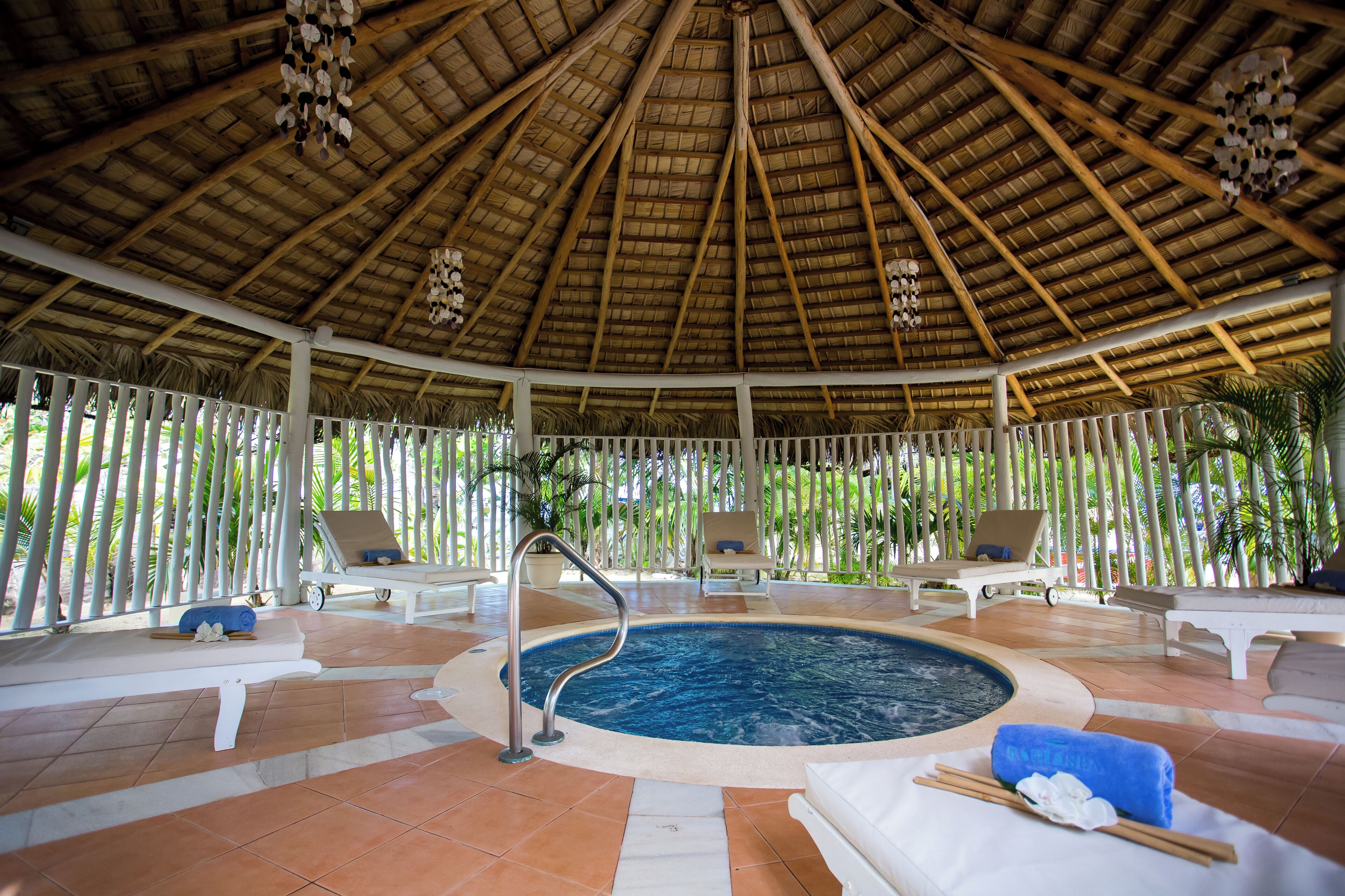 A covered pool area with lounge chairs, a hot tub, and a thatched roof.