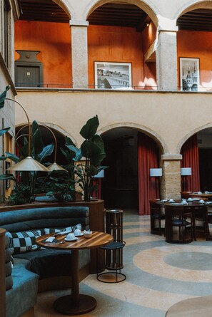 A spacious lobby with a high ceiling, featuring a central seating area with a sofa and tables, surrounded by large potted plants and a patterned floor.