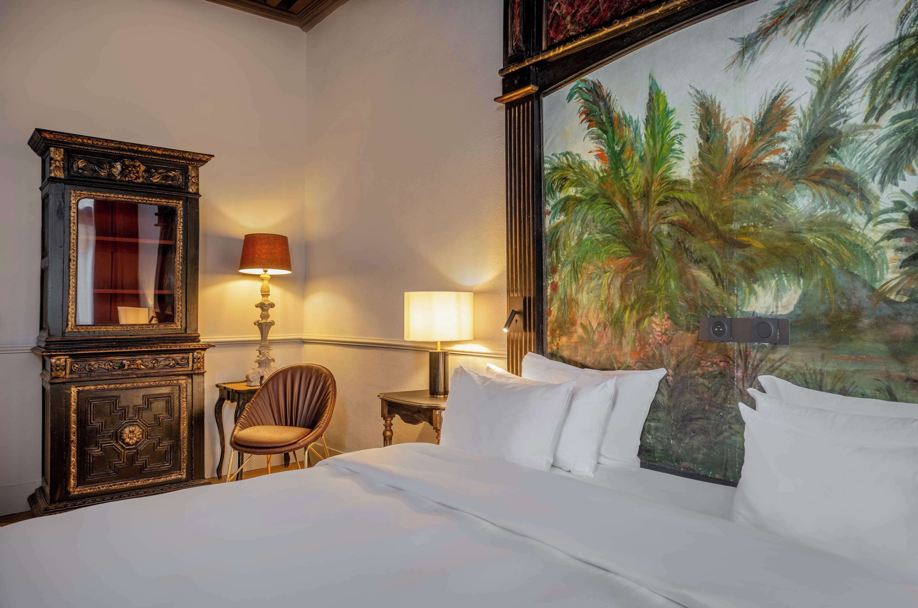 A hotel room with a bed, two lamps, a chair, a desk, and a large painting of palm trees.