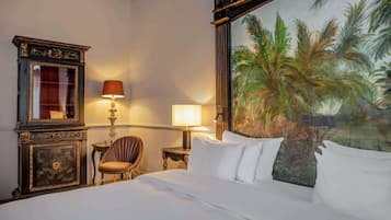 A hotel room with a bed, two lamps, a chair, a desk, and a large painting of palm trees.
