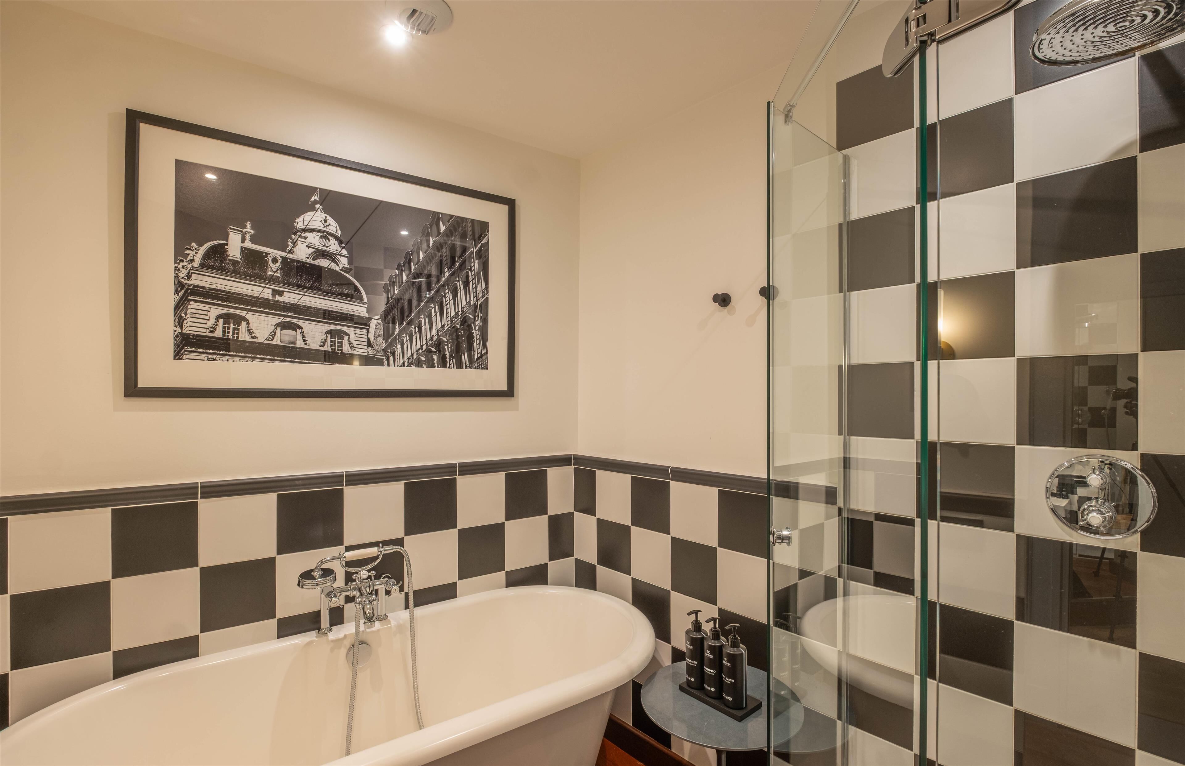 Une salle de bain avec un mur recouvert de carreaux noirs et blancs formant un motif à carreaux, une photographie en noir et blanc encadrée, une baignoire sur pieds et un espace de douche délimité par des parois en verre.