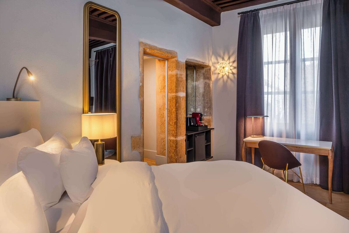 A hotel room with a bed, a desk, a chair, a mirror, and a window with curtains.