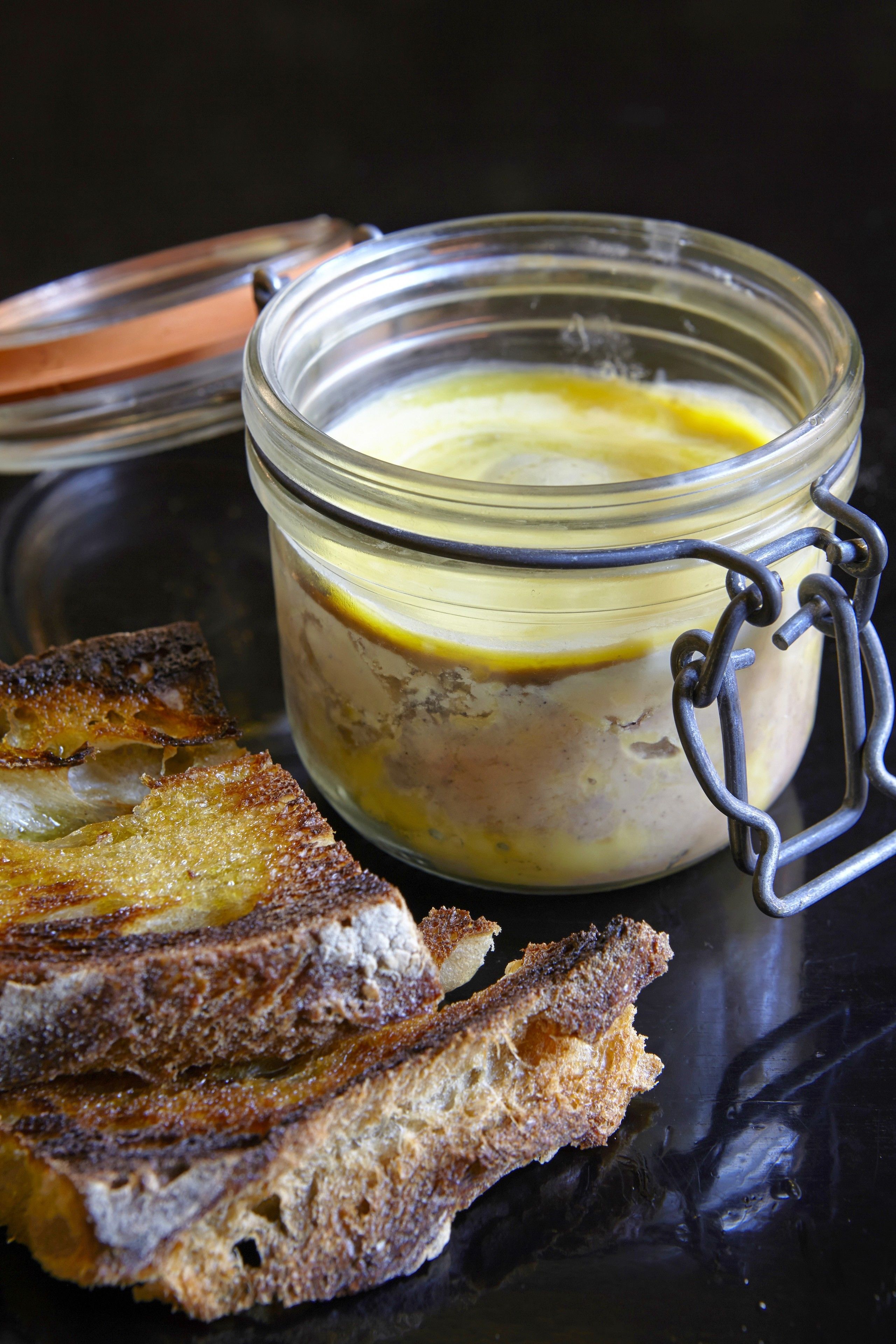 A glass jar with a metal clasp, containing a yellowish substance, and a piece of toasted bread beside it.