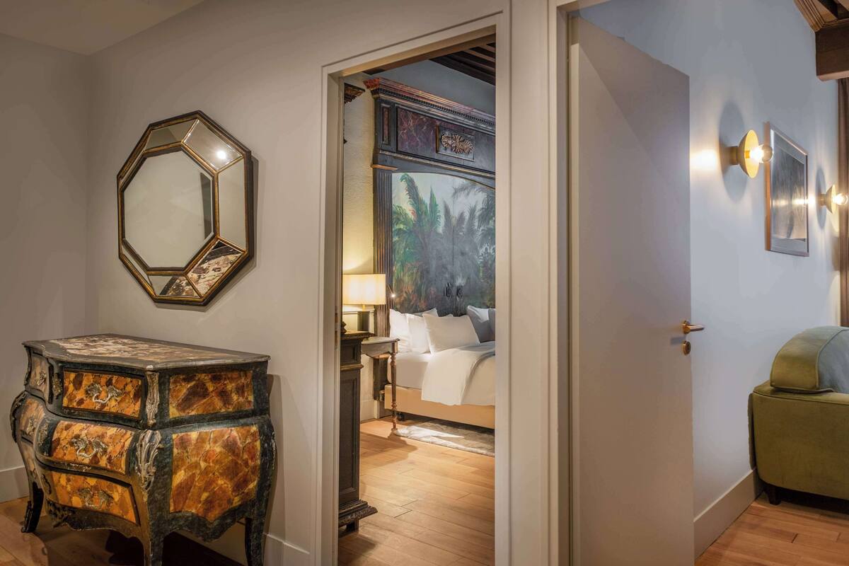 A room with a wooden floor, a green armchair, a bed with white linens, a nightstand with a lamp, and a hexagonal mirror on the wall.