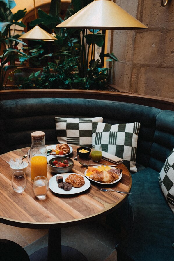 A cozy dining area with a round wooden table set for breakfast, featuring croissants, fruit, and a bottle of orange juice. A checkered pillow and a lamp are on a green sofa in the background.