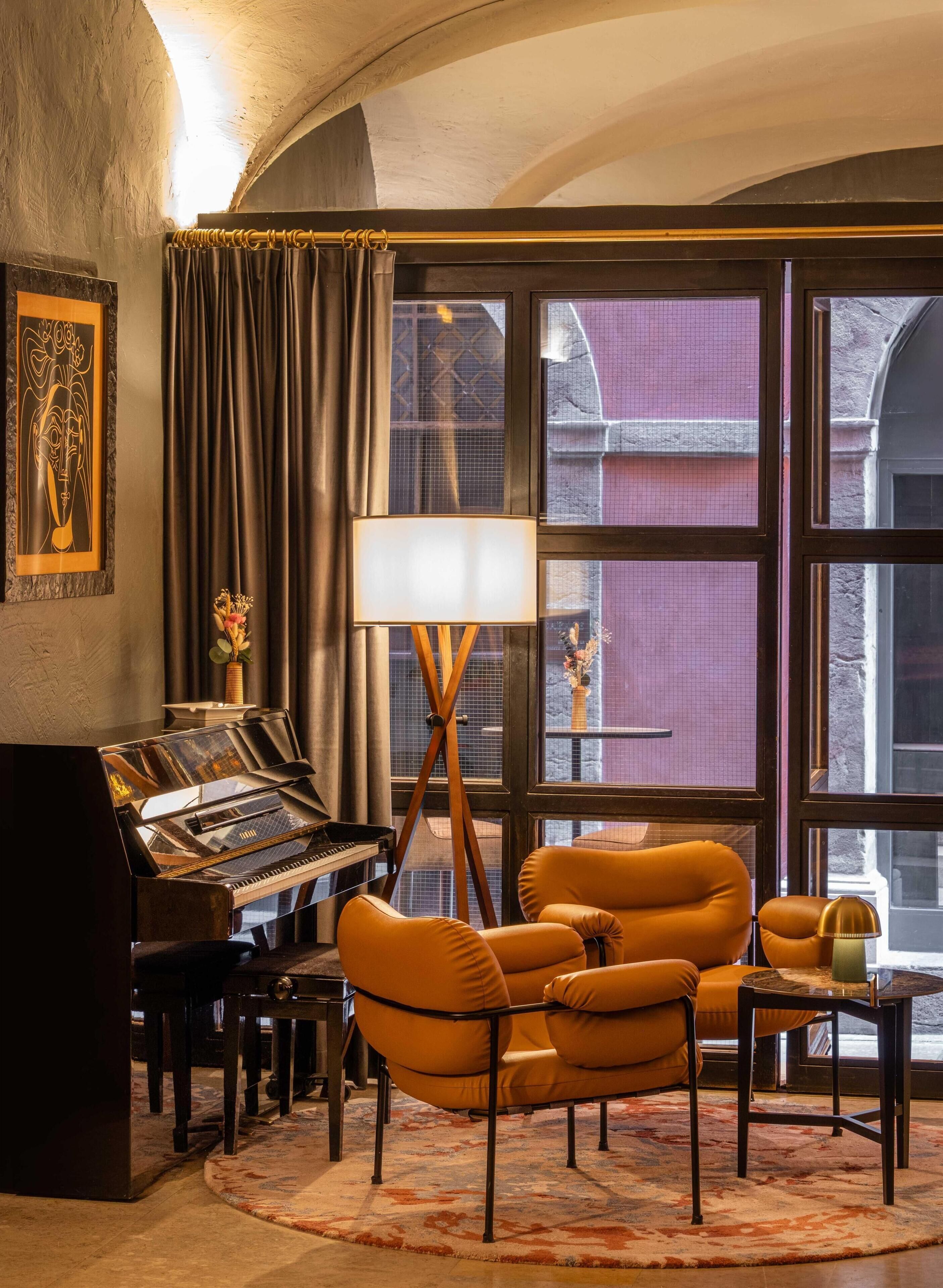 Une pièce comprenant un piano à queue, deux fauteuils confortables, une table d’appoint, une lampe et une grande fenêtre donnant sur des immeubles.