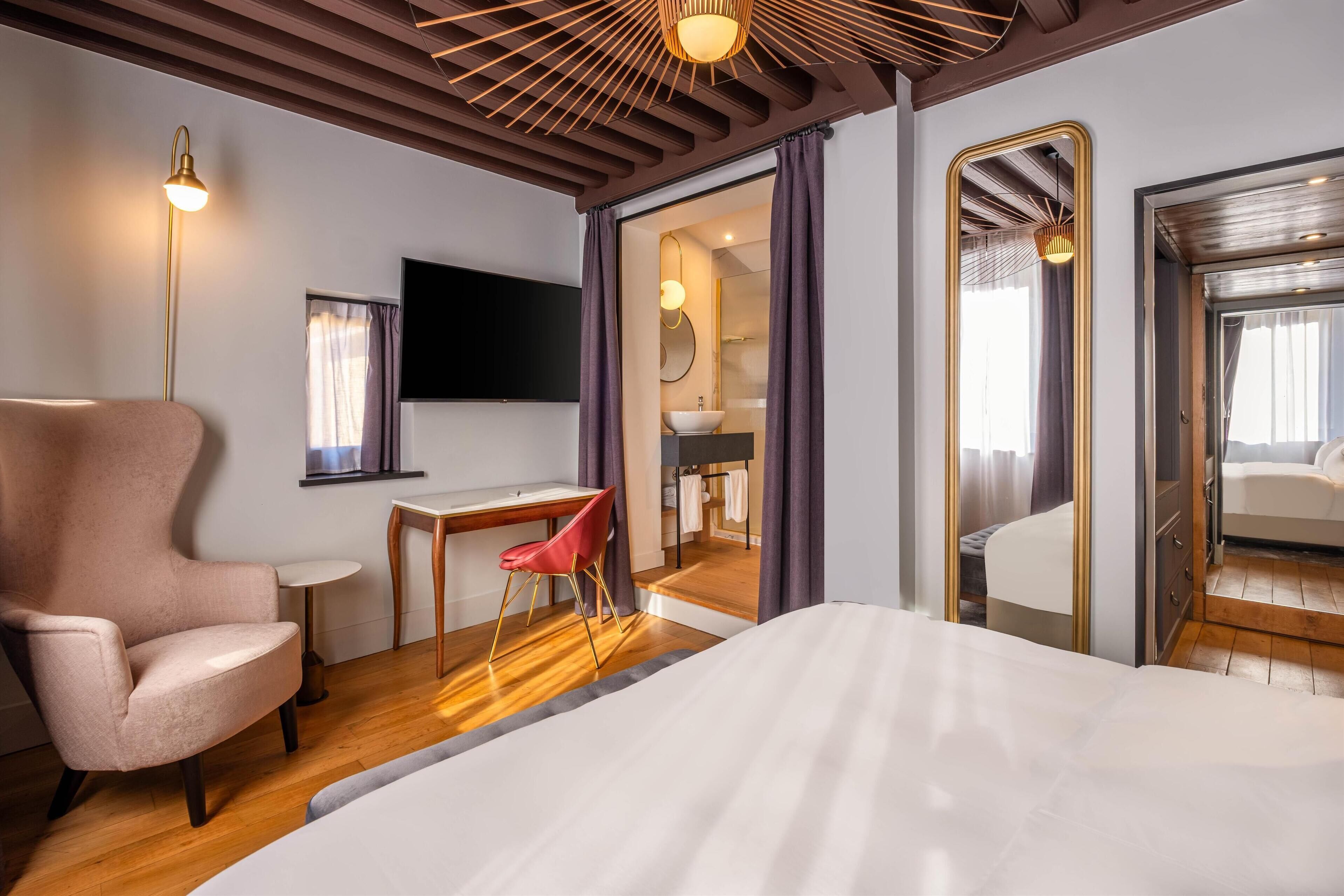 Quarto de hotel moderno com uma cama grande, uma escrivaninha com cadeira, televisão e banheiro visível através de uma porta aberta.