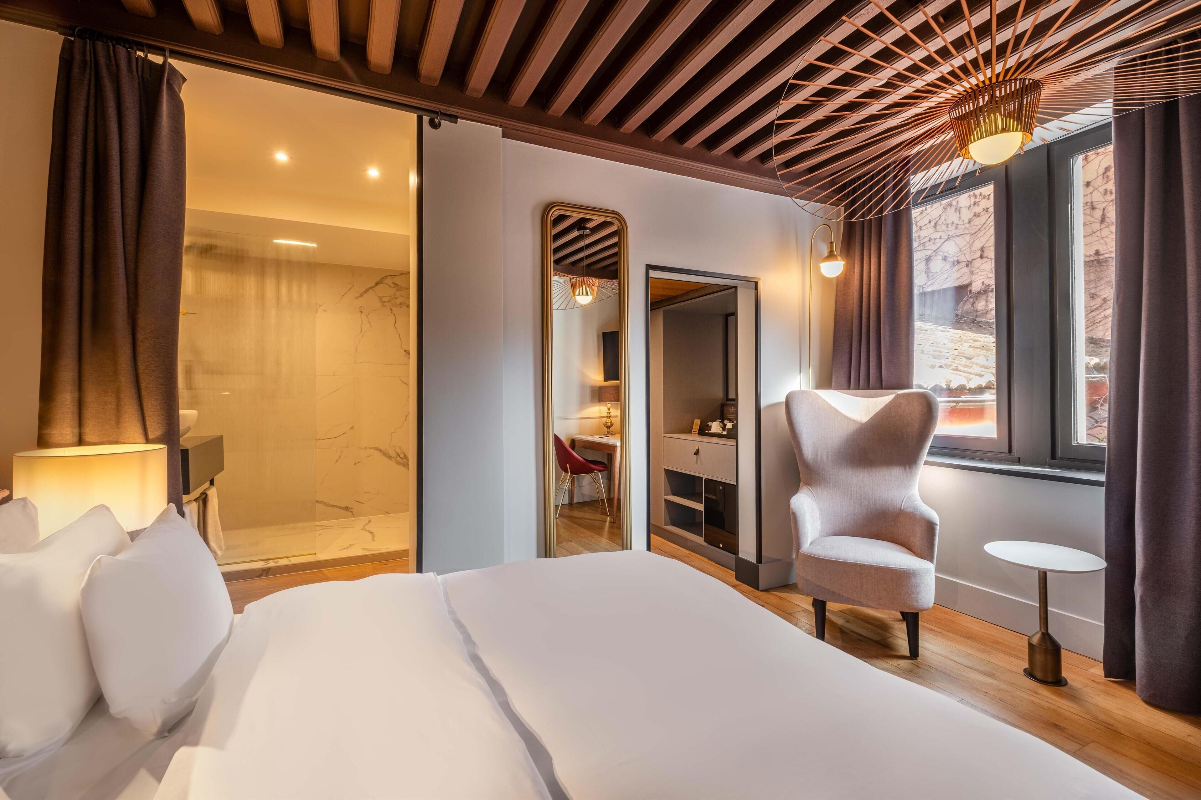 Habitación de hotel moderna con una cama grande, una zona de estar con una silla, una pequeña mesa, un vestidor y un baño con ducha de vidrio.