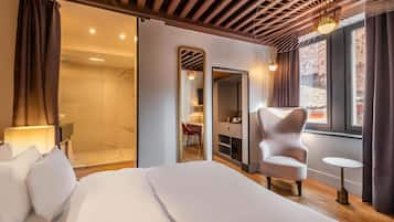 Habitación de hotel moderna con una cama grande, una zona de estar con una silla, una pequeña mesa, un vestidor y un baño con ducha de vidrio.