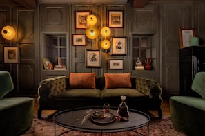 A cozy living room with a green sofa, two orange cushions, a round coffee table with a decanter and glasses, and framed pictures on the wall.