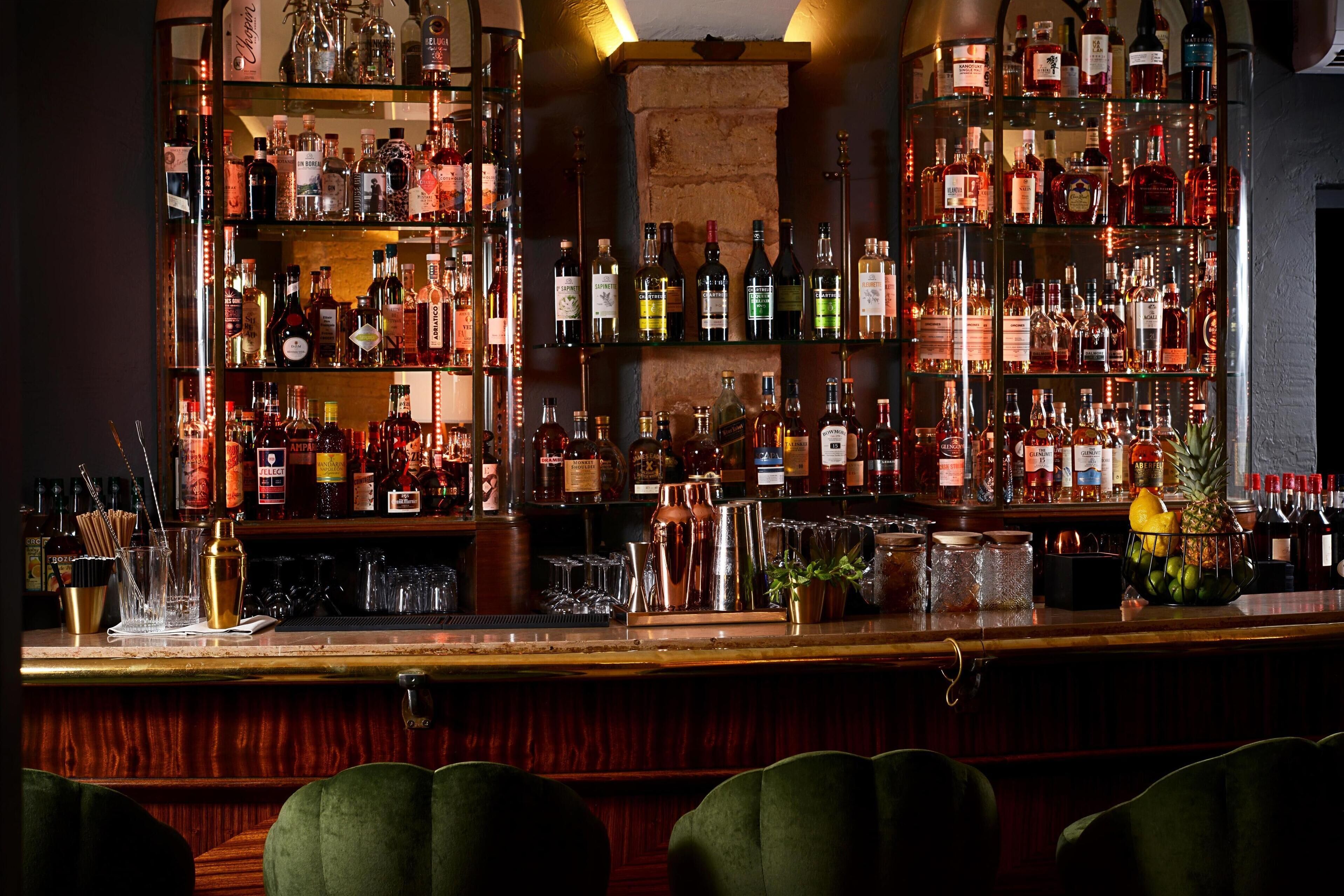 A bar with multiple shelves of liquor bottles, a bar counter with bar tools, and a fruit basket.