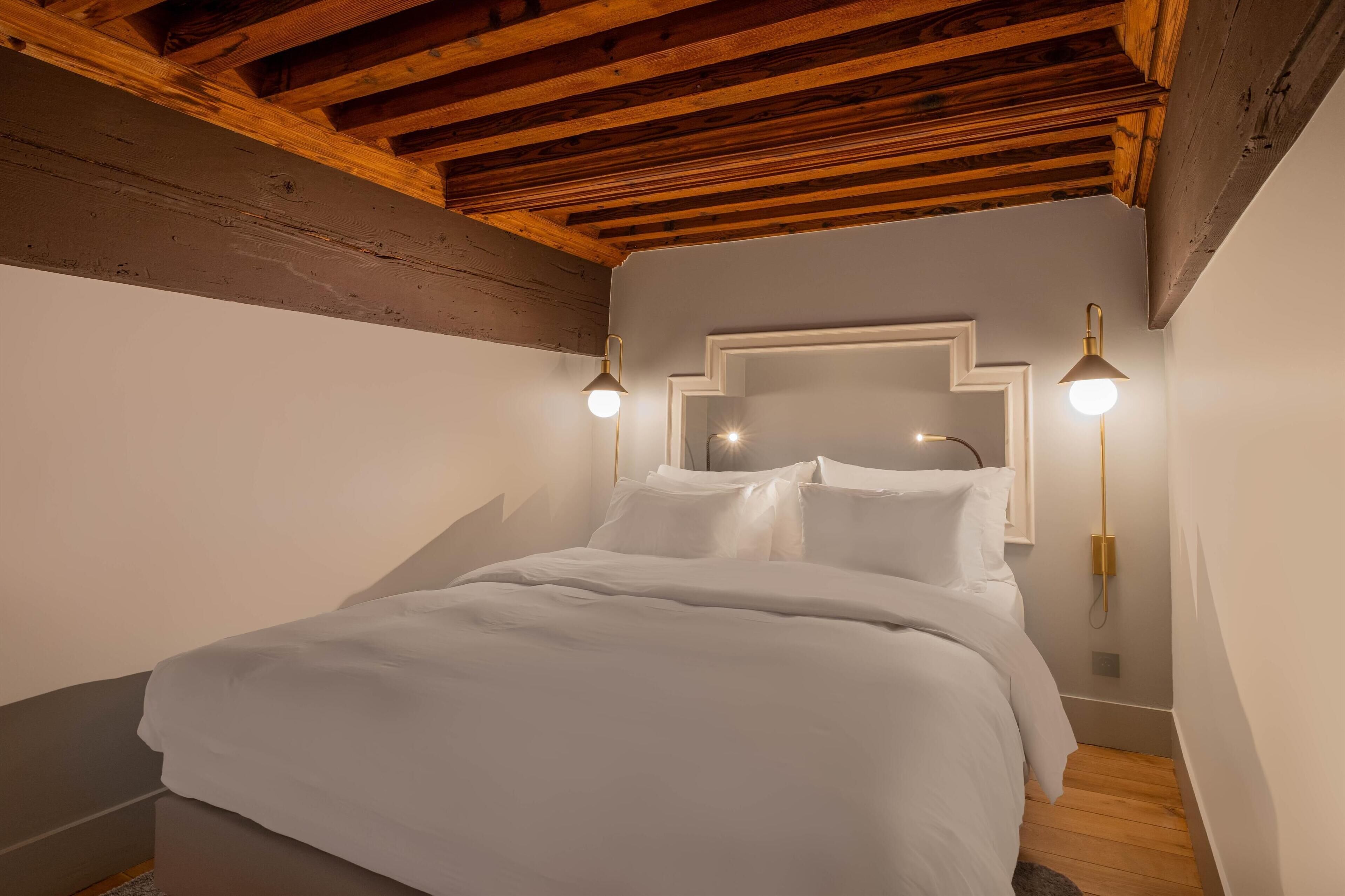 Un dormitorio con techo de madera, una cama con ropa de cama blanca y dos lámparas fijadas a la pared.