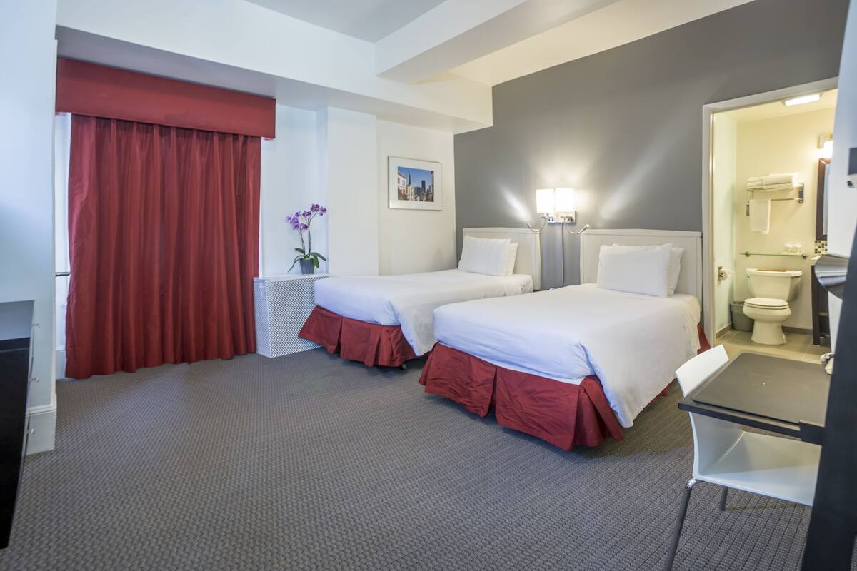 A hotel room with two beds, a desk, a chair, a window with red curtains, a small table with a vase of flowers, and a bathroom visible through an open door.