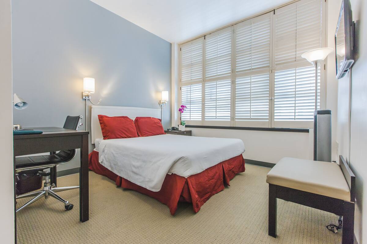A hotel room with a bed, desk, chair, and large window with blinds.