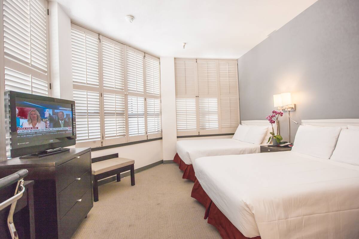 A hotel room with two beds, a TV on a dresser, and large windows with blinds.