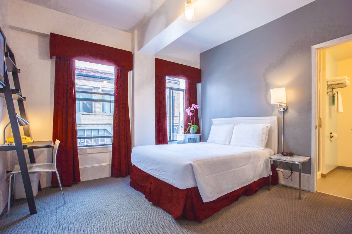 A hotel room with a bed, a desk, a chair, a window with curtains, a bedside table with a lamp, and a door.
