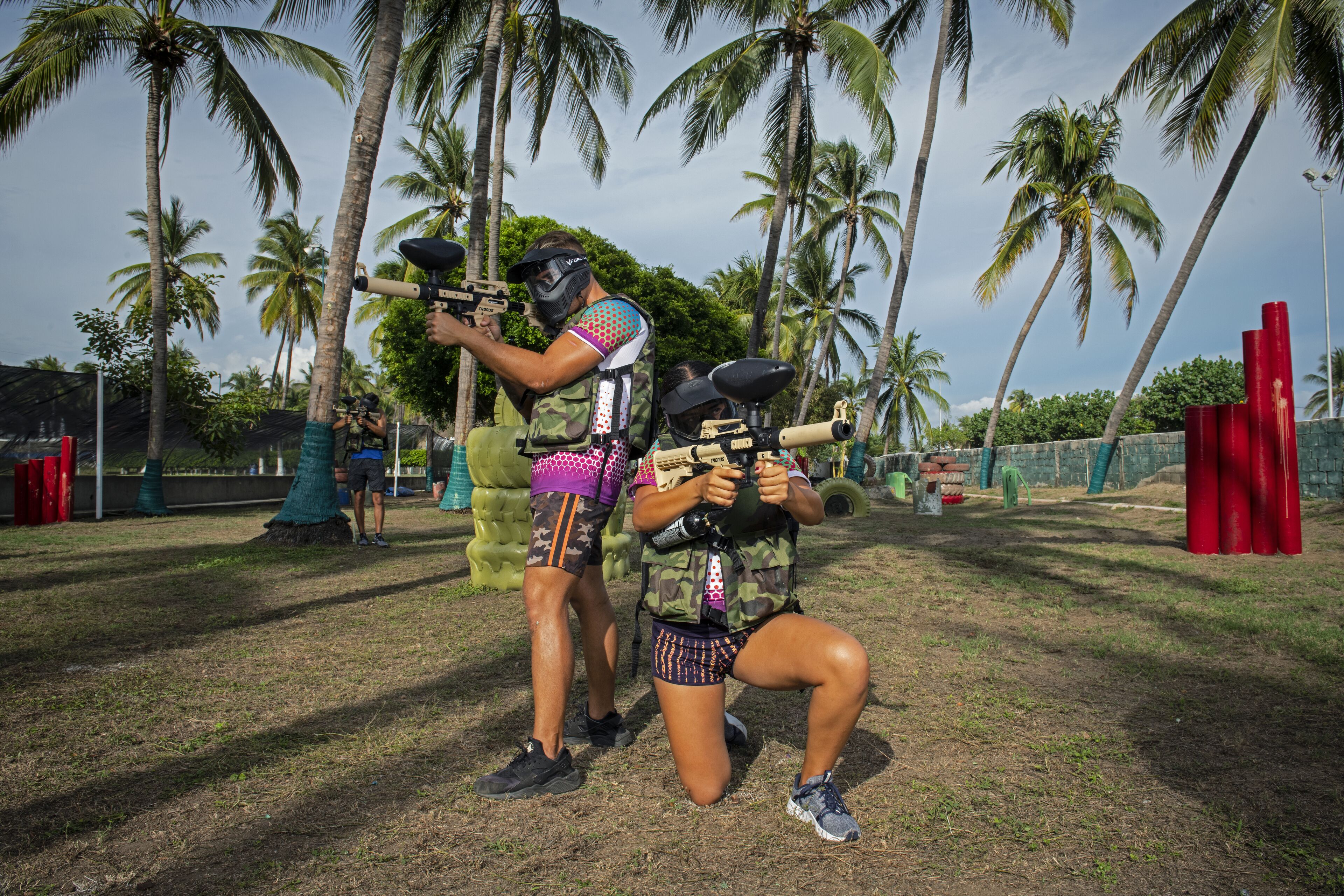 Two people in camouflage attire with paintball gear, standing in a grassy area with palm trees in the background.