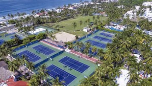 Vista aèria d'una zona costanera amb diverses pistes de tennis envoltades de palmeres i edificis.