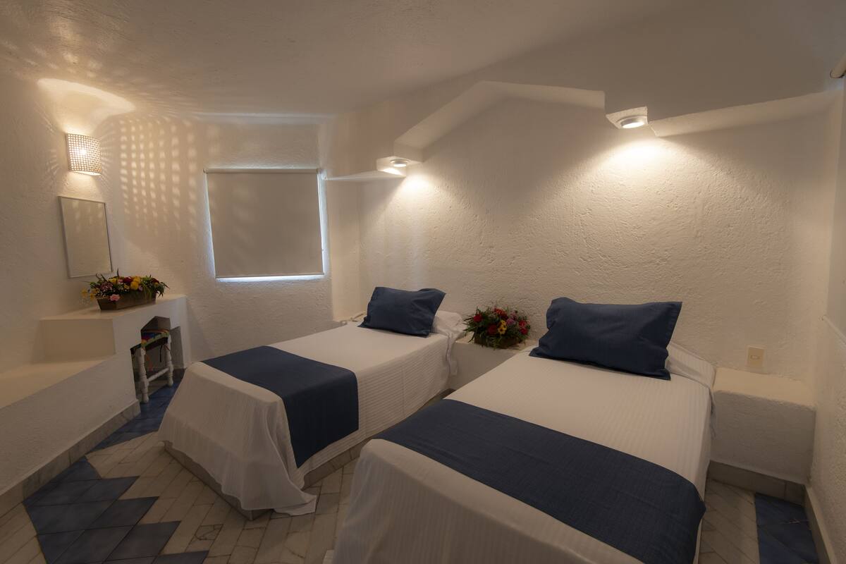 A bedroom with two beds, a small window, a nightstand, a mirror, and a wall-mounted light fixture.
