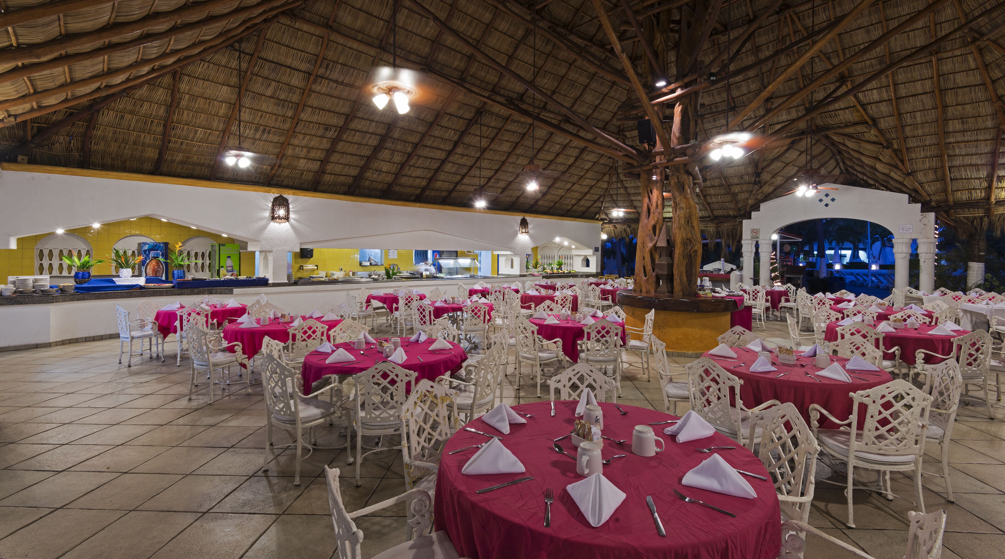 Un restaurant avec des tables rondes recouvertes de nappes rouges, des chaises blanches et un plafond en chaume.