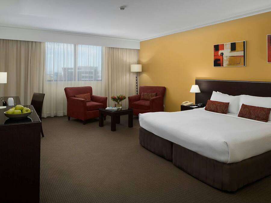Quarto de hotel com uma cama grande, duas poltronas vermelhas, uma mesinha com um vaso, uma escrivaninha com uma fruteira e uma televisão.