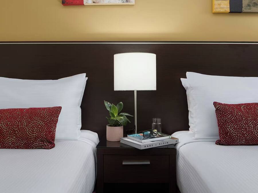 Une chambre d’hôtel avec deux lits, du linge de lit blanc, des coussins à motifs rouges, une table de chevet avec une lampe, une petite plante en pot et un livre.