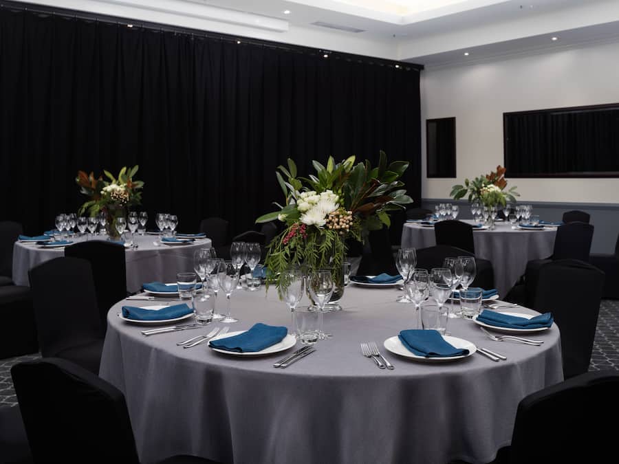 A conference room with round tables, each set with plates, cutlery, and glassware, and decorated with floral centerpieces.