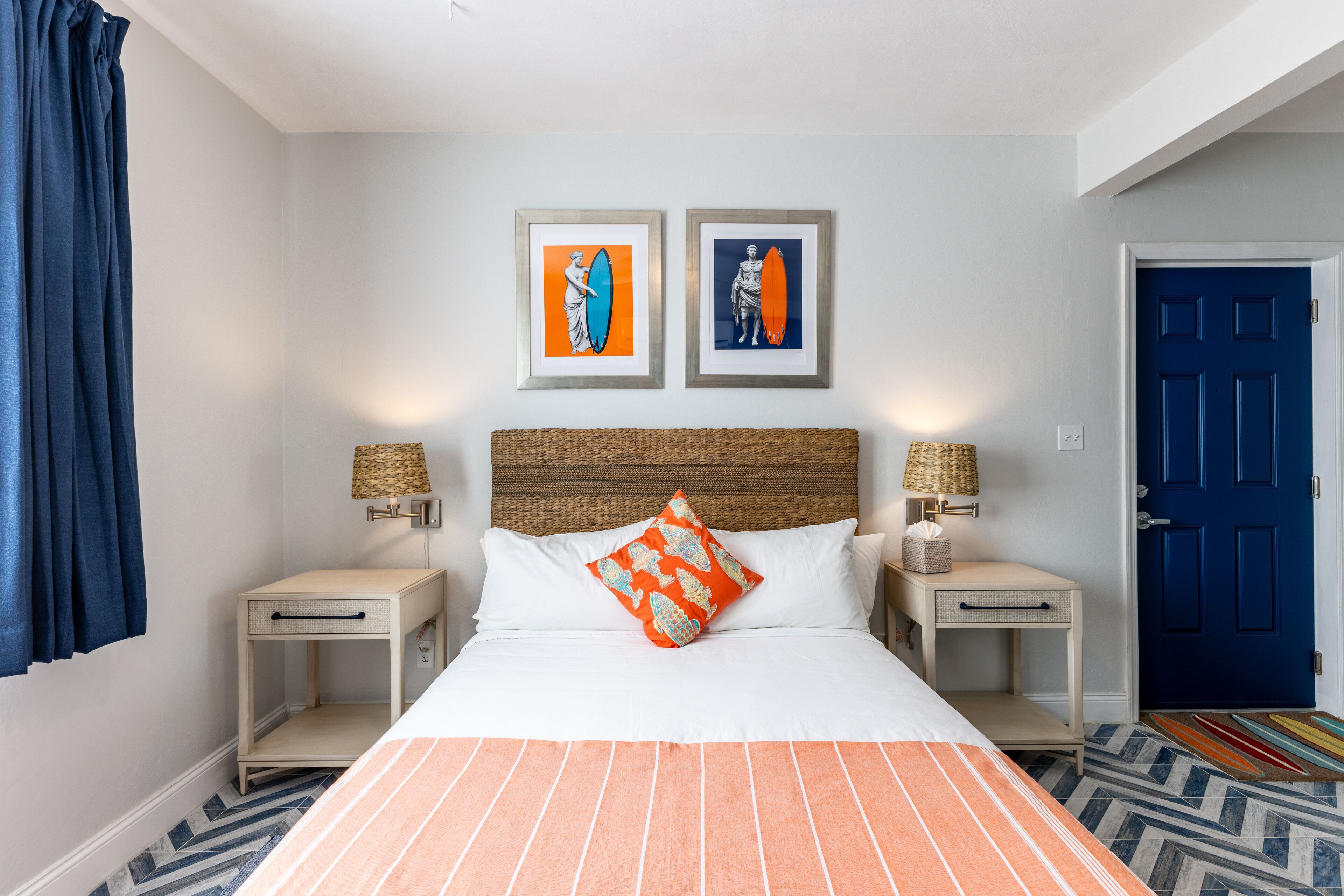 Una habitación de hotel con una cama, dos mesitas de noche, una puerta azul y dos cuadros en la pared.