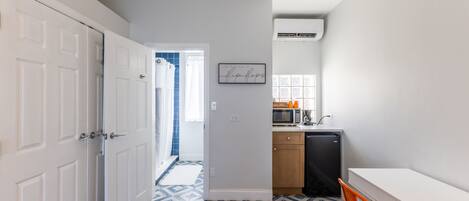 Un pasillo con piso de estampado geométrico, una puerta blanca, una zona de cocina con microondas y una pequeña mesa con una silla.