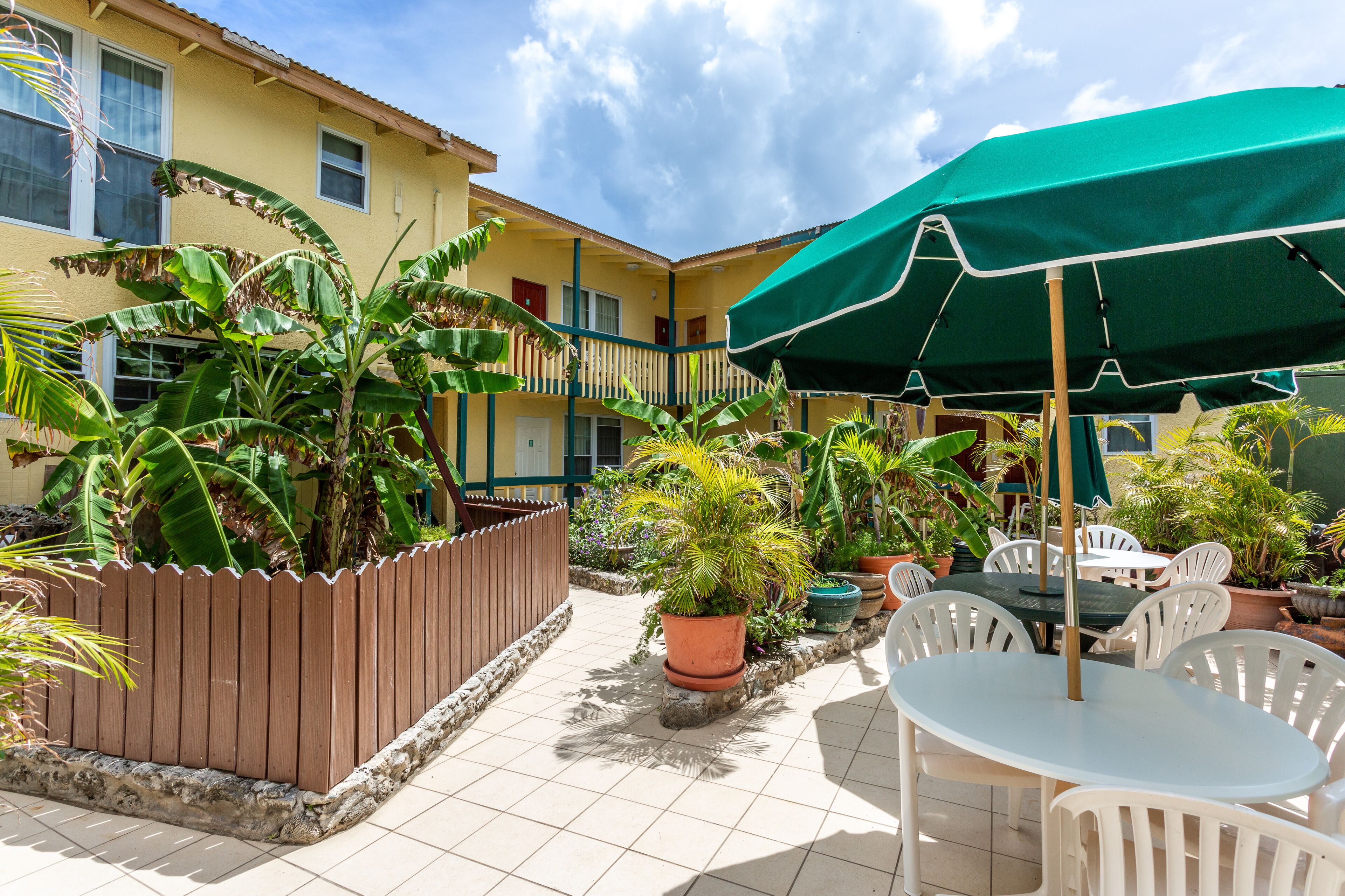 A patio area with a table and chairs under a green umbrella, surrounded by potted plants and a wooden fence.