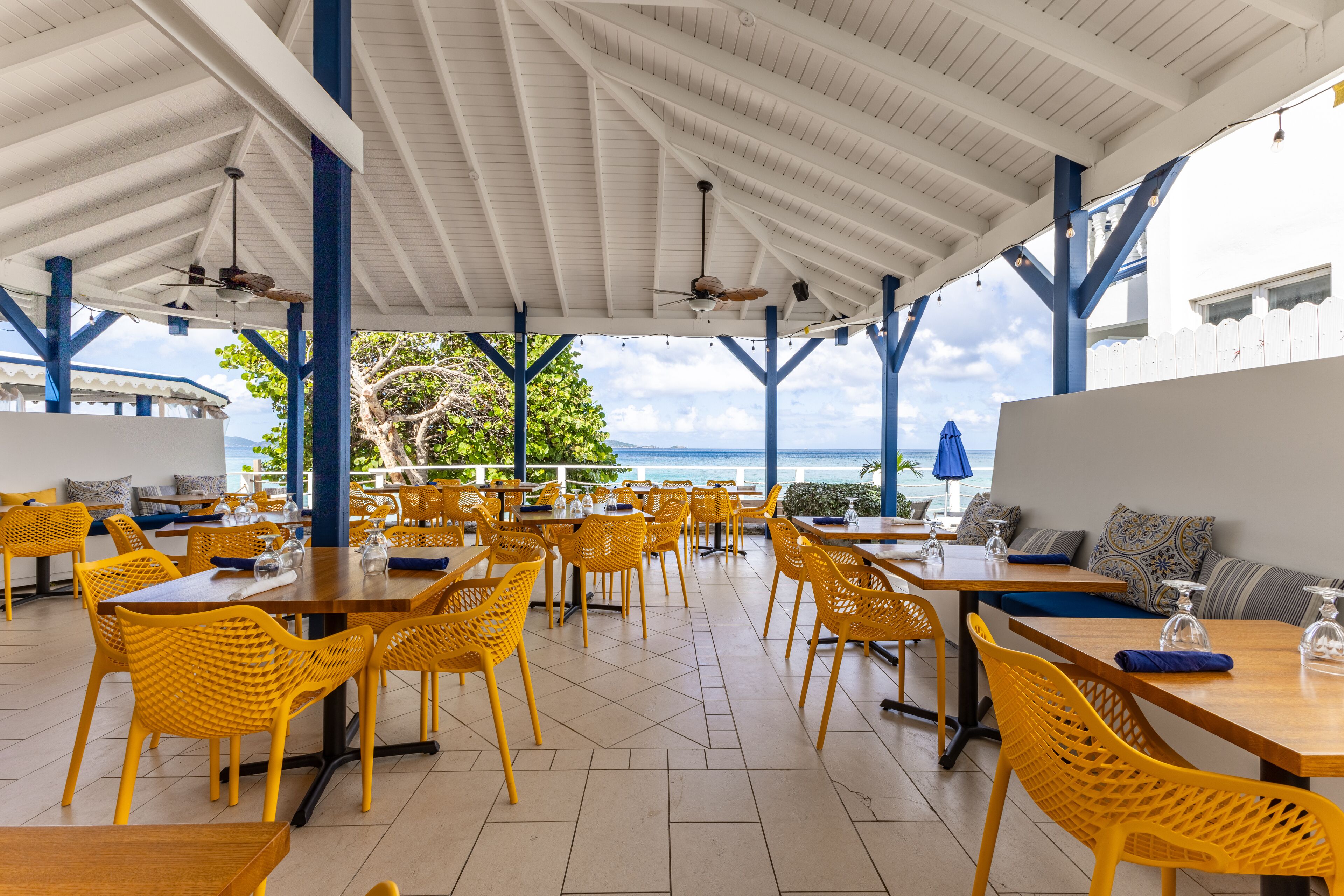 A covered outdoor dining area with yellow chairs and tables, a white ceiling, and a view of the ocean.