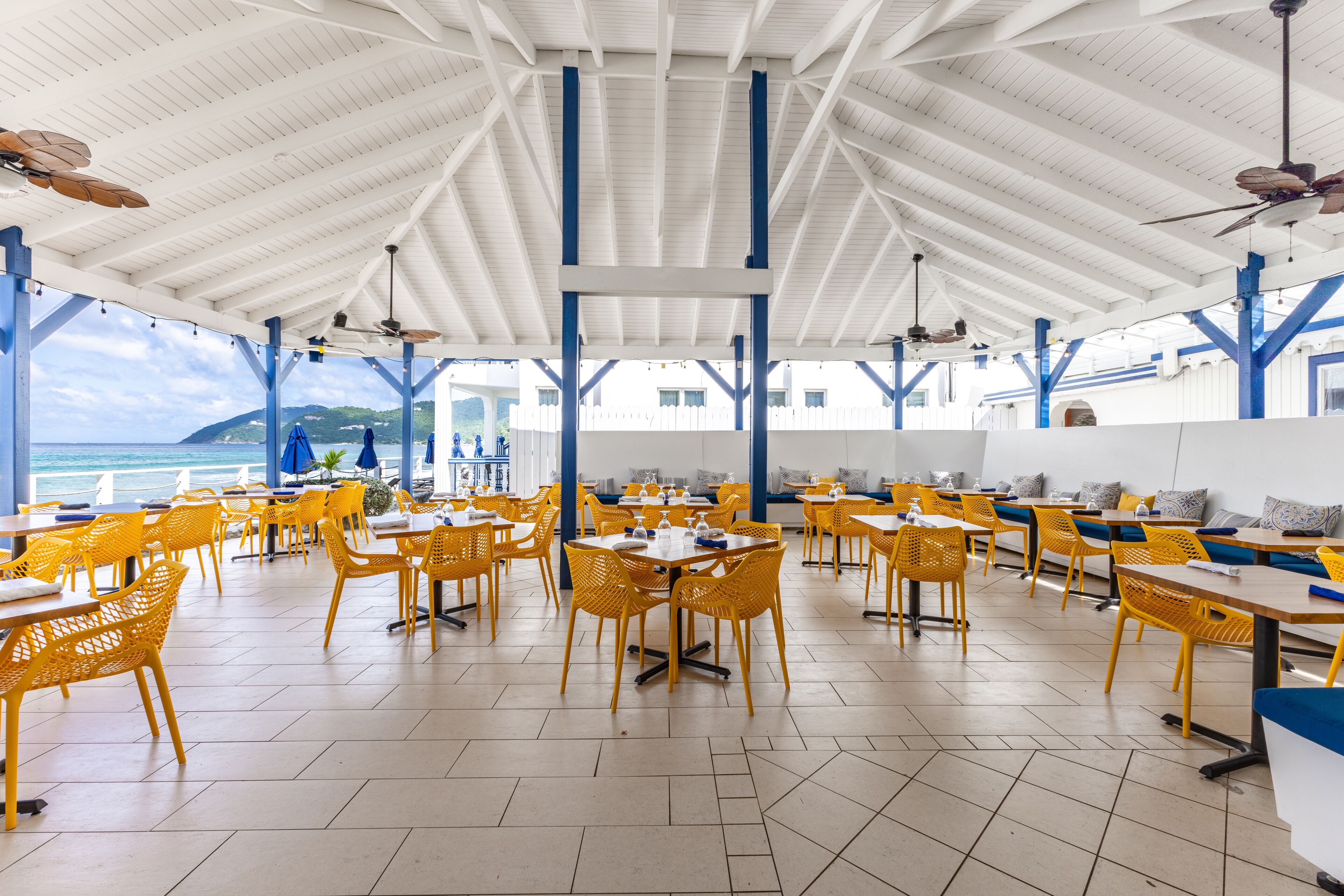 A spacious outdoor dining area with yellow chairs and tables, white ceiling, and blue structural beams.