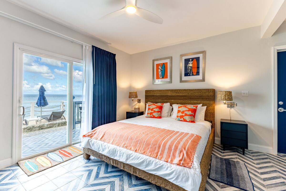 Quarto de hotel com cama, mesa de cabeceira, luminária, vista para a praia e uma porta.