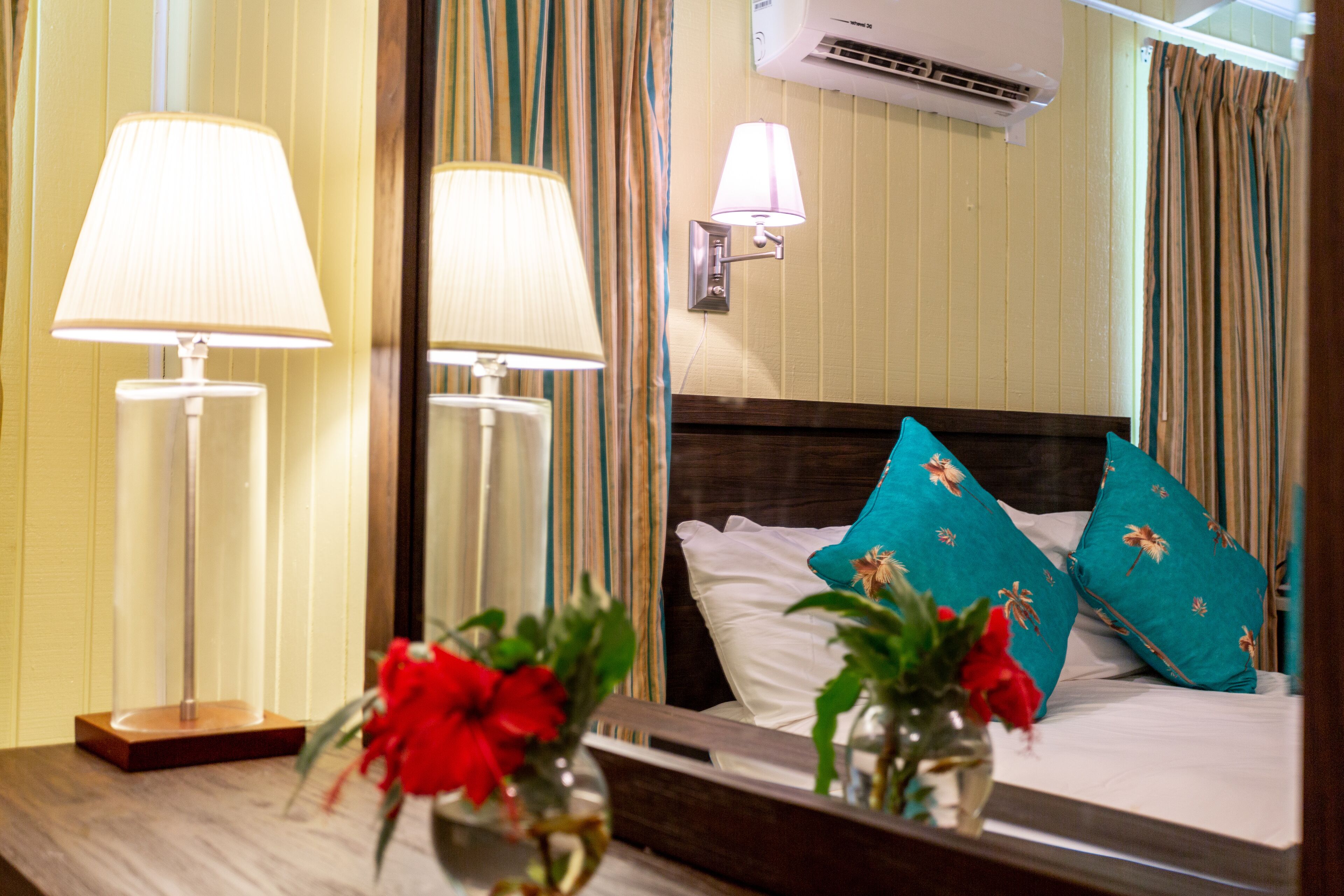 Una habitación de hotel con una cama, dos lámparas de noche, un espejo y un jarrón con flores rojas.