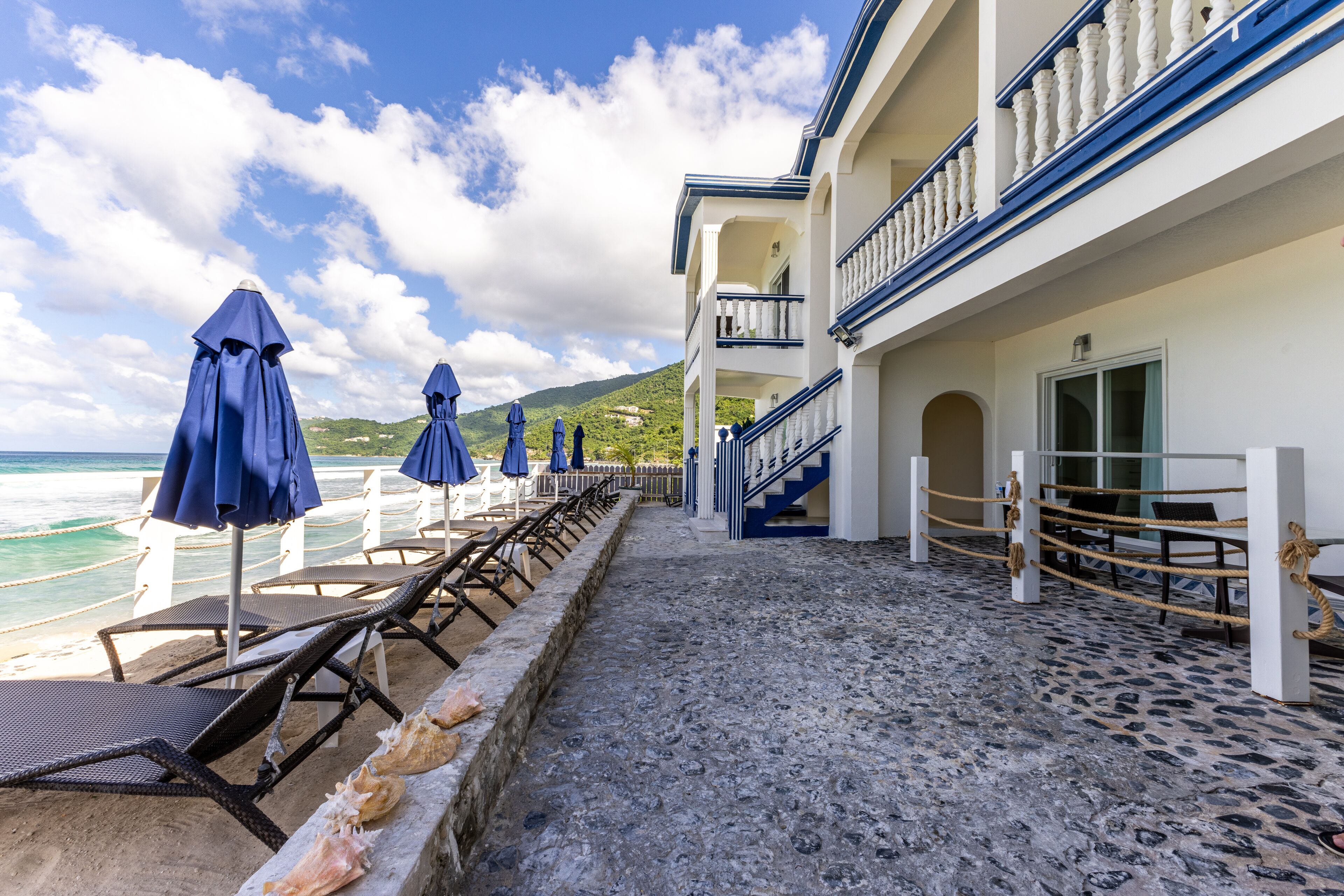 Ein Strandhotel mit Liegestühlen, Sonnenschirmen, einem gepflasterten Weg und einem Balkon mit Geländer.
