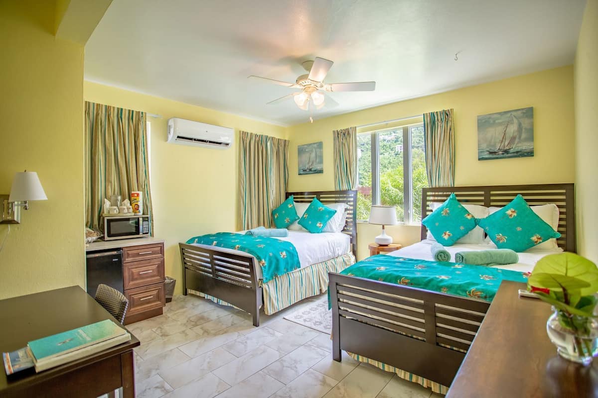 Quarto de hotel com duas camas, uma escrivaninha, micro-ondas, ventoinha no teto e janela com cortinas.